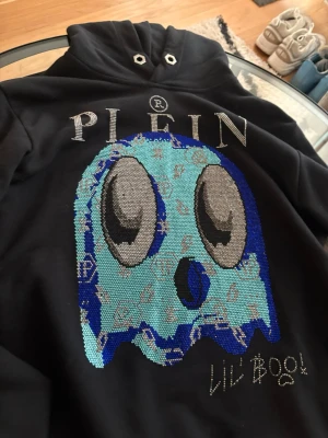 Philipp Plein hoodie med stenar  - Snygg svart hoodie från Philipp Plein med stor, glittrig Pac-Man-inspirerad spökdesign i blått och silver på bröstet. Märkets logga syns tydligt ovanför motivet. Hoodien har huva med metallöljetter och känns riktigt exklusiv och unik.