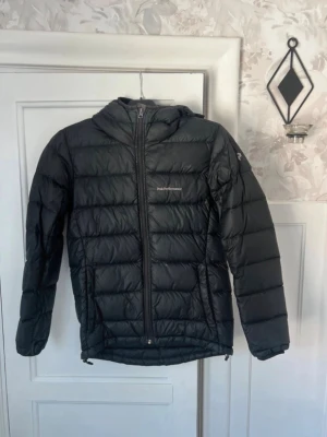 Svart dunjacka från Peak Performance - Snygg svart dunjacka från Peak Performance med huva och quiltad design. Jackan har dragkedja framtill och långa ärmar. Perfekt för kyliga dagar och enkel att matcha med olika outfits.