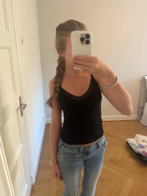 Svart linnetopp med spetskant - Snygg svart linnetopp med tunna axelband och dekorativ spetskant vid ringningen. Toppen har en figurnära passform och är perfekt att matcha med jeans eller kjol. Materialet känns mjukt och stretchigt, vilket gör den bekväm att bära.