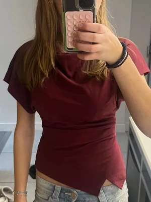Vinröd topp med asymmetrisk nederkant - Snygg vinröd topp med korta ärmar och unik asymmetrisk nederkant. Toppen har en tight passform och är tillverkad i ett mjukt, stretchigt material som sitter skönt på kroppen. Perfekt att styla med både jeans och kjol.