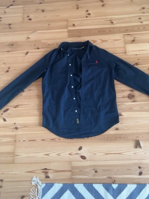Mörkblå skjorta från Ralph Lauren - Klassisk mörkblå skjorta från Ralph Lauren med röd broderad logga på bröstet. Skjortan har långa ärmar, knappar framtill och en tidlös passform. Perfekt för dig som gillar stilrena plagg med ikonisk detalj. Säljer tröjan då den är för stor, inte använd utan bara provad.          Pris kan diskuteras!