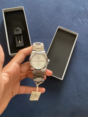 Silverfärgad klocka i helt nytt skick. - Stilren klocka i silver med klassiskt länkarmband i metall. Urtavlan är vit med tydliga markeringar och datumvisning. Klockan levereras i originalask och verktyg för att justera länken och även extra batteri medföljer.