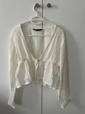 Vit blus från Zara - Supersöt vit blus från Zara med djup V-ringning, knytband i midjan och volangdetaljer. Tillverkad i ett tunt, lätt och lite skrynkligt tyg som ger en avslappnad boho-vibe. Långärmad modell med ballongärmar och spetskant längs ringningen.