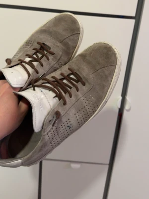 Gråa sneakers i mocka från Tod's - Snygga gråa sneakers från Tod's i mjuk mocka med vita detaljer och bruna skosnören. Skorna har perforerade sidor för extra ventilation och en vit sula med metalliska detaljer baktill. Perfekt för dig som gillar stilrena och klassiska sneakers. Nypris 5000kr mitt pris 500kr