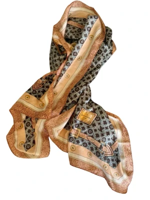 Mönstrad sjal från Louis Vuitton - Snygg sjal från Louis Vuitton med klassiskt monogrammönster i svart, beige och brunt. Sjalen har detaljerade kanter med knappliknande motiv och en lyxig känsla i materialet, troligen siden. Perfekt accessoar för att lyfta din outfit. 55*155 cm. 