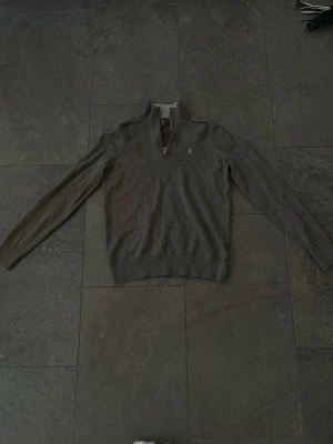 Grå stickad tröja med dragkedja Morris - Snygg grå stickad tröja från Morris i 100% lammull. Tröjan har halv dragkedja med läderdetalj, hög krage och diskret broderad logga på bröstet. Perfekt för lager-på-lager och en stilren look.