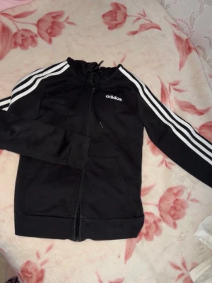 Svart Adidas hoodie med dragkedja - Säljer en äkta svart kofta från Adidas med dragkedja framtill och klassiska vita ränder längs ärmarna. Tröjan har huva med dragsko, två fickor och Adidas-logga på bröstet. Perfekt för en sportig och avslappnad stil. Passar även för er som har storlek S.