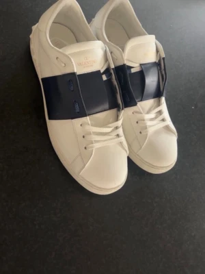 Valentino vita sneakers med blå detalj - Snygga vita sneakers från Valentino med bred mörkblå remsa över mitten. Klassisk låg modell med rund tå och vita snören. Tillverkade i slätt läder och har diskret Valentino-logga på plösen. Perfekta för dig som gillar stilrena och exklusiva sneakers.