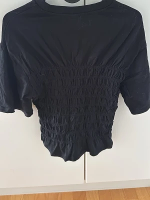 Svart croppad topp med smock - Säljer en svart croppad topp med smockad midja och korta ärmar. Toppen har rund halsringning och är gjord i mjukt material som sitter bekvämt mot kroppen. Perfekt till jeans eller kjol för en trendig look.