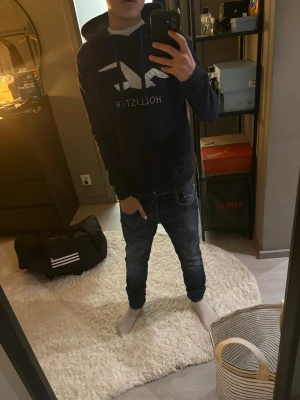 Mörkblå hoodie från Hollister - Snygg mörkblå hoodie från Hollister med stor vit logga och text på bröstet. Klassisk känguruficka framtill och justerbar huva med dragsko. Tillverkad i mjukt material som passar perfekt till jeans eller joggers.