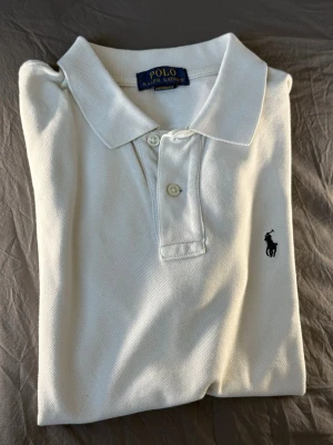 Vit pikétröja från Ralph Lauren - Klassisk vit pikétröja från Ralph Lauren med blå broderad logga på bröstet. Tröjan har krage, knappar framtill och är tillverkad i bomull för en bekväm känsla. Perfekt till jeans eller shorts för en clean look.