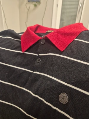 Randig långärmad polotopp med röd krage från K-boxing - Snygg långärmad polotopp från Lacoste med svart bas, vita horisontella ränder och en kontrasterande röd krage. Tröjan har knappar framtill och en diskret logga på bröstet. Perfekt för dig som gillar klassisk men ändå färgstark stil.