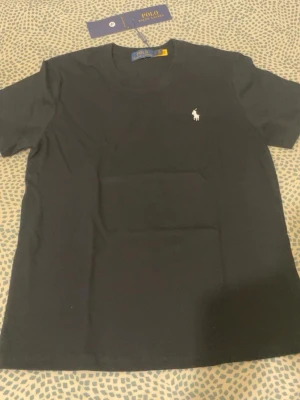 Svart t-shirt från Polo Ralph Lauren - Klassisk svart t-shirt från Polo Ralph Lauren med rund halsringning och den ikoniska vita logon broderad på bröstet. Tillverkad i mjuk bomull för en skön känsla och perfekt passform. Enkel och stilren design som funkar till allt.