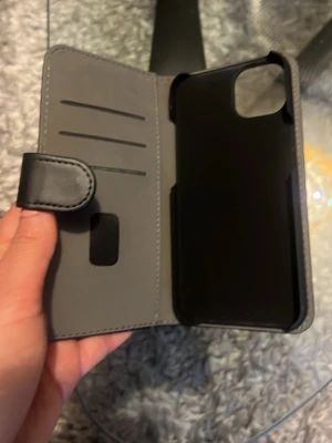 iPhone 13 Svart plånboksfodral till mobil  - Snyggt svart plånboksfodral till mobil med plats för kort och magnetlås. Fodralet har en stilren design och är gjort i konstläder med ett hårt plastskal på insidan för extra skydd. Perfekt för dig som vill ha både mobil och kort på samma ställe.