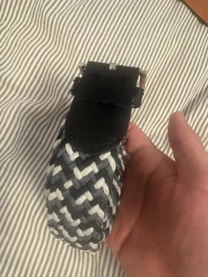 Randigt flätat bälte i svart och vitt - Snyggt flätat bälte med randigt mönster i svart och vitt. Bältet har ett klassiskt spänne i metall och detaljer i svart skinn. Perfekt accessoar för att lyfta din outfit.