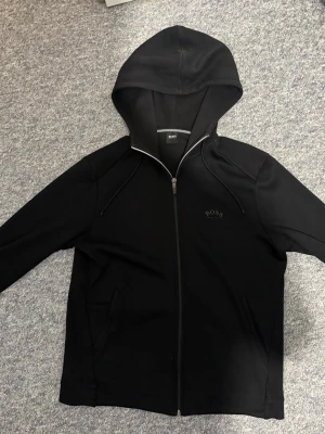 Svart hoodie BOSS - Snygg svart hoodie från BOSS med dragkedja framtill och justerbar huva med snören. Diskret BOSS-logga på bröstet och två fickor på sidorna. Tillverkad i mjukt material som känns bekvämt mot huden. Perfekt för en avslappnad och stilren look.