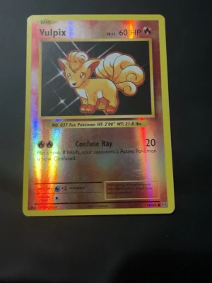 Vulpix holografiskt Pokémonkort - Säljer ett holografiskt Vulpix Pokémonkort med gul ram och glittrande yta. Kortet har motiv av Vulpix, nivå 11, 60 HP och attacken Confuse Ray. Baksidan har klassiskt blått Pokémon-mönster med Pokéboll. Perfekt för samlare eller Pokémonfans.