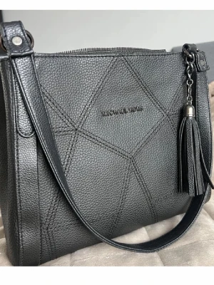 Svart handväska från Michael Kors - Snygg svart handväska från Michael Kors i strukturerat läder med dekorativa sömmar och en stor tofs som detalj. Väskan har axelrem och dragkedja upptill. Perfekt för dig som vill ha en stilren och trendig accessoar.