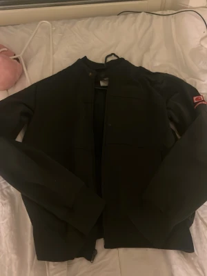 Svart Carhartt jacka med patch - Säljer en svart jacka från Carhartt med röd och vit patch på ärmen. Jackan har en stilren design med hög krage och dragkedja framtill. Perfekt för dig som gillar streetwear och enkel stil.