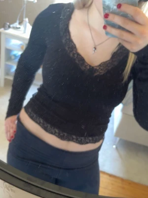 Svart långärmad topp med spets - Snygg svart långärmad topp med v-ringning och spetsdetaljer längs halsringningen och nederkanten. Modellen är figurnära och toppen är croppad vilket ger en trendig look. Perfekt att matcha med höga jeans eller kjol.