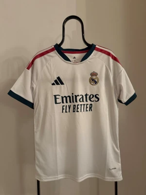 Real Madrid matchtröja Adidas - Snygg vit Real Madrid matchtröja från Adidas med klubbmärke på bröstet och sponsortryck 'Emirates Fly Better'. Tröjan har mörkblå detaljer vid ärmslut och krage samt röda ränder på axlarna. Tillverkad i lätt och ventilerande material, perfekt för fotbollsträning.