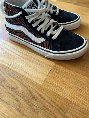 Vans Sk8-Hi med zebramönster - Säljer ett par Vans Sk8-Hi sneakers med svart mocka och brunt zebramönster på sidorna. Klassisk vit sula och vit sidestripe, samt vita skosnören. Perfekta för dig som vill sticka ut med coola detaljer och streetwear-vibe.