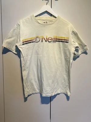 Ljus Gul O'Neill t-shirt med retrotryck - Säljer en gul t-shirt från O'Neill i 100% bomull. T-shirten har ett retroinspirerat tryck med gula och bruna ränder samt O'Neill-loggan över bröstet. Klassisk passform och rund hals, perfekt för en avslappnad stil.