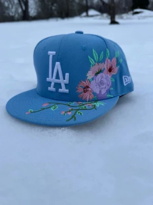 Blå LA New Era keps med broderat blommönster  - Snygg blå keps från New Era med LA-logga framtill i vitt och broderade blommor i rosa, lila och grönt på både skärmen och sidan. Klassisk flatbrätte och 59FIFTY-modell. Perfekt för dig som vill sticka ut med en unik accessoar.pris kan absolut diskuteras 