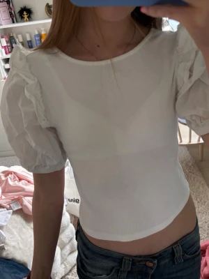 Vit croppad topp med puffärm - Söt vit croppad topp med puffiga kortärmar och volangdetaljer på axlarna. Toppen har rund halsringning fram och fyrkantig rygg. Perfekt för dig som gillar en feminin och trendig stil.