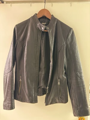 Svart skinnjacka från DKNY - Snygg svart skinnjacka från DKNY med ståkrage och dragkedja framtill. Jackan har quiltade detaljer på axlar och ärmar samt två fickor med dragkedja. Perfekt för en cool och stilren look.