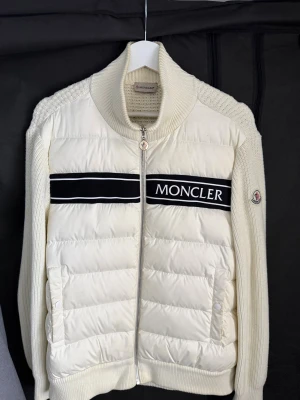 Vit Moncler stickad dunjacka - Säljer en snygg vit dunjacka från Moncler med stickad struktur och ribbade detaljer. Jackan har dragkedja med Moncler-logga och är långärmad. Materialet är en mix av ull och polyester med dunfyllning, perfekt för kyliga dagar. Stilren och clean look.