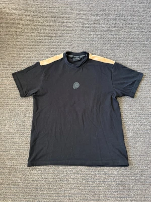 T-shirt  - Snygg svart t-shirt från Bywear med beige detaljer över axlarna och ett diskret emblem på bröstet. Klassisk rund hals och korta ärmar. Perfekt till jeans eller shorts för en clean och stilren look.