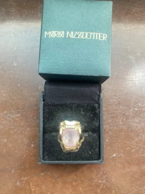 Byte Maria Nilsdotter Jaw Ring - Unik ring från Maria Nilsdotter i guldtonad metall med en stor, ljusrosa sten i mitten. Den är 16 mm och tyvärr för liten för mig, byter mot en 17 mm. Aldrig använd. 