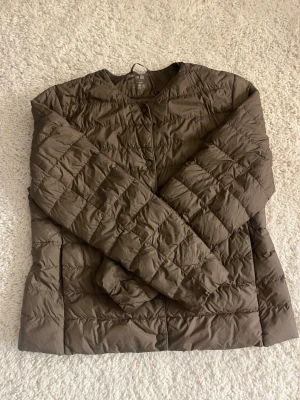 Brun dunjacka från Uniqlo - Lätt och stilren brun dunjacka från Uniqlo med rund halsringning och tryckknappar framtill. Jackan har ett quiltat mönster och är perfekt för kalla vinterdagar. Enkel design utan huva och med långa ärmar. Passar dig som gillar minimalistisk stil.