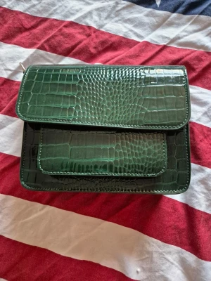 Grön croco-mönstrad handväska - Snygg grön handväska i croco-mönstrat konstläder med två fickor framtill och magnetlås. Väskan har en fyrkantig form och glansig finish som ger en trendig look. Perfekt för dig som vill sticka ut med en cool accessoar.