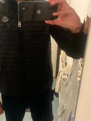 Svart dunväst från Moncler - Säljer en svart dunväst från Moncler med quiltad design och dragkedja framtill. Västen har Moncler-logga på bröstet och två sidofickor med dragkedja. Perfekt för lager-på-lager och riktigt snygg till hoodie eller tröja.