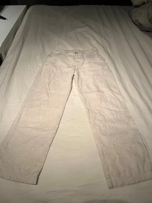 Beiga linnebyxor med raka ben - Säljer ett par beiga linnebyxor med raka ben och hög midja. Byxorna har knapp och dragkedja framtill samt resår i midjan baktill för extra komfort. Tillverkade i ett luftigt linnematerial som passar perfekt till sommaren.