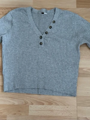 Grå ribbstickad tröja med knappar - Snygg grå ribbstickad tröja med v-ringning och dekorativa knappar längs halsringningen. Tröjan har långa ärmar och en mjuk, stretchig känsla. Perfekt för lager på lager och enkel att matcha med jeans eller kjol. Storlek och tyglappar bortklippt.