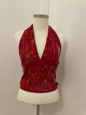 Röd halterneck topp med paljetter - Snygg röd halterneck topp med djup v-ringning och dekorativa paljetter samt broderade blommor framtill. Toppen är ärmlös och har öppen rygg, perfekt för dig som vill sticka ut. Materialet känns mjukt och stretchigt, vilket ger en bekväm passform. Vissa paljetter fattas.