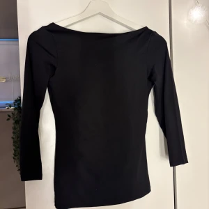 Svart topp med 3/4-ärm och vid ringning - En stilren svart topp med trekvartsärmar och bred, djup ringning bak. Figurnära passform och mjukt, stretchigt material som ger en clean look. Perfekt att matcha med jeans eller kjol för en enkel men snygg outfit.