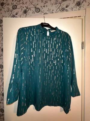 Grön blus med metallicdetaljer - Stilren grön blus från KappAhl med långa ärmar och hög hals. Blusen har ett mönster av vertikala metallicdetaljer i silver som ger ett snyggt skimmer. Knäppning i nacken med knapp och liten öppning. Perfekt för dig som gillar att sticka ut.