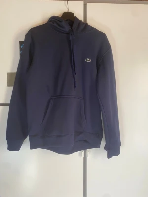 Mörkblå hoodie från Lacoste - Snygg mörkblå hoodie från Lacoste med klassisk krokodillogga på bröstet. Hoodien har huva med dragsko och en stor magficka framtill. Tillverkad i mjukt material som känns skönt mot huden. Perfekt för en avslappnad och stilren look.