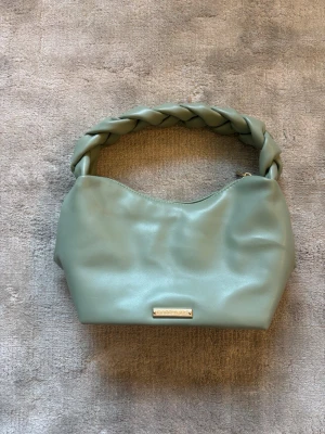 Grön handväska med flätat handtag - Ljusgrön handväska från Steve Madden i mjukt konstläder. Bredd: 26 cm. Djup: 14 cm. Höjd: 15 cm. 