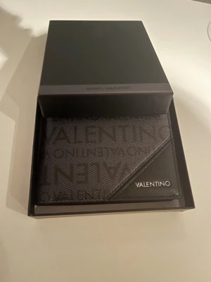 Svart plånbok från Mario Valentino - Snygg svart plånbok från Mario Valentino med logotypdetaljer i tyg och läder. Plånboken har flera kortfack och ett större fack för sedlar. Stilig design med VALENTINO-tryck på framsidan och metallogo. Perfekt för dig som vill ha en trendig och praktisk accessoar.