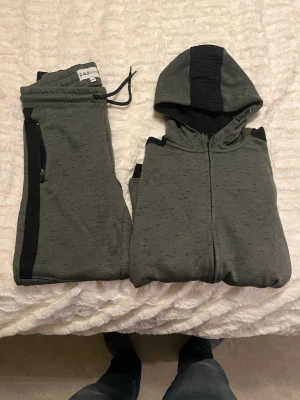 Grönmelerad loungewear-set med huva - Säljer ett grönmelerat loungewear-set från D.O.D med svarta detaljer. Setet består av en zip-hoodie med huva och två fickor med dragkedja samt matchande byxor med dragsko och svarta sidopaneler. Perfekt för chill dagar hemma.