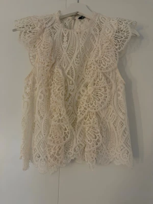 Vit spetsblus med volangdetaljer - Supersöt vit blus i spets med volangdetaljer längs framsidan och axlarna. Ärmlös modell med rund halsringning och vackert mönstrad spets. 