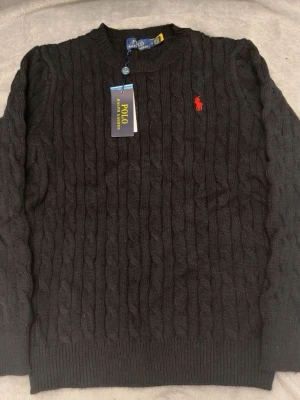 Svart kabelstickad tröja Polo Ralph Lauren - Kabelstickad svart tröja från Polo Ralph Lauren med rund halsringning och röd broderad logga på bröstet. Klassisk design med ribbade muddar vid ärmslut och nederkant. Perfekt för en stilren och tidlös look.