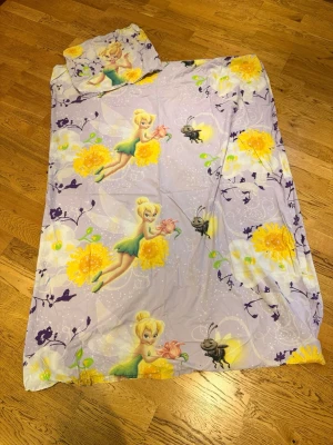 Lila Tingeling påslakanset - Säljer ett påslakanset med motiv av Tingeling från Disney. Setet är lila med stora gula blommor, fjärilar och figurer från filmen. Materialet är mjukt bomullstyg och både påslakan och örngott ingår. Perfekt för dig som gillar Disney och vill ha ett färgglatt sängset.