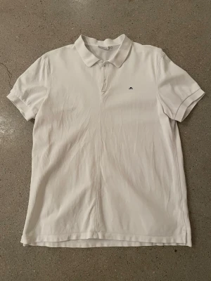 Vit pikétröja från J.Lindeberg - Klassisk vit pikétröja från J.Lindeberg med liten logga på bröstet. Tröjan har krage, knappar framtill och korta ärmar. Perfekt till både jeans och shorts för en clean look.