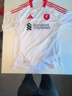Vit Liverpool matchtröja Adidas - Snygg vit Liverpool FC matchtröja från Adidas med röda detaljer och klubbmärke på bröstet. Tröjan har tre röda ränder på axlarna, svart Standard Chartered-tryck och är tillverkad i lätt, ventilerande funktionsmaterial. Perfekt för fotboll eller träning, eller bara ha ute 